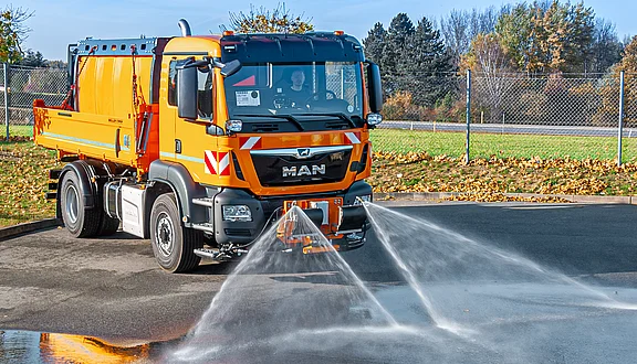 Ein orangefarbener MAN-Lkw steht auf einem Asphaltplatz. Aus der Vorderseite sprühen zwei Schwemmdüsen, die den Boden reinigen. Im Hintergrund sind hohe Bäume und ein Zaun sichtbar, während der Himmel klar ist.