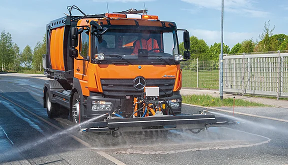 Ein orangefarbener MAN mit einem Schwemmbalken fährt auf einer Straße. Das Fahrzeug sprüht mit großem Druck das Wasser auf die Fahrbahn um diese zu reinigen. Im Hintergrund sind Bäume und ein Zaun sichtbar.