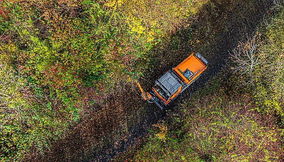 Ein Kommunalfahrzeug nutzt einen Lichtraumprofilschneider um einen Weg wieder begehbar zu machen. Er befindet sich auf einem unbefestigten Weg, umgeben von üppigem, grünem Laub und Bäumen in herbstlichen Farben.