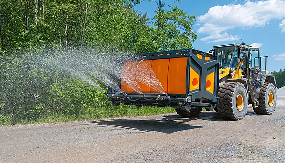 Ein großer Radlader mit einem orangefarbenen Tank auf der Ladegabel sprüht Wasser auf eine unbefestigte Straße. Der Wagen fährt durch eine landschaftliche Umgebung mit Bäumen im Hintergrund.