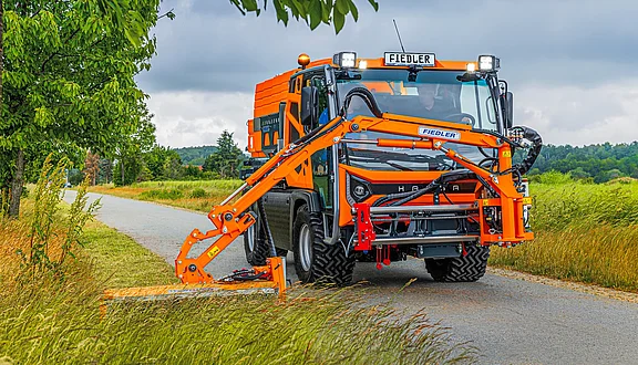 Ein kleiner orangefarbener Lkw steht an einem Landweg, umgeben von Bäumen und hohem Gras. Er verfügt über einen Frontausleger, der für verschiedene Arbeiten eingesetzt werden kann. Aktuell ist er mit einem Flachmähkopf ausgestattet und mäht den Straßenrand.