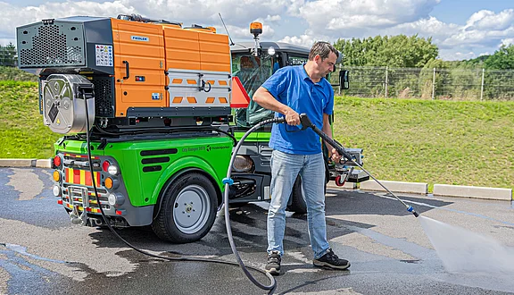 Ein Mann bedient einen Hochdruckreiniger, der an einem kleinen, grünen Arbeitsfahrzeug befestigt ist. Er sprüht Wasser auf den Boden, während im Hintergrund eine bewachsene Fläche und ein blauer Himmel zu sehen sind.