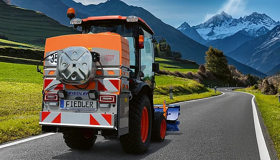Ein Traktor steht auf einer kurvenreichen Landstraße, umgeben von grünen Wiesen und Bergen im Hintergrund. Die Maschine hat orangefarbenen Tank mit einem Bürstensystem an der Vorderseite. Der Schriftzug "FIEDLER" ist deutlich sichtbar.