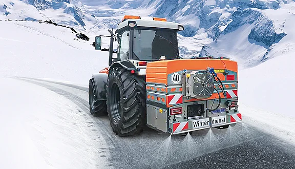 Winterdienst-Traktor mit Fiedler Solesprühanlage im Einsatz: Effiziente Glatteisbekämpfung durch Soleverteilung auf verschneiter Gebirgsstraße. Die Heck-Sprühanlage ist in verschiedenen Tank-Volumina erhältlich.
