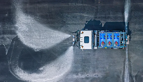 Ein MAN reinigt mit den Fiedler Schwemmdüsen die asphaltierte Fläche. Der LKW hat ein blaues Tanksystem. Der steuerbare Sprühstrahlen verteilen sich gleichmäßig auf dem Boden, um Schmutz zu entfernen.