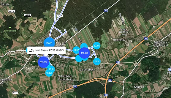 Eine Kartenansicht zeigt einen Ort mit verschiedenen blauen Kreisen, in denen die ausgebrachte Menge von Streugut auf den Straßen dargestellt ist. Ein Punkt beschreibt das Fahrzeug mit montierten Anbaugerät.