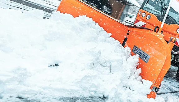 Ein orangefarbener Schneepflug räumt frischen Schnee von einer Straße. Es schneit stark, und der Schneepflug ist aktiv dabei, die Schneemassen zu beseitigen.