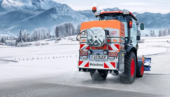 Traktor mit Fiedler Winterdienst-Kombination: Kommunalschlepper im Einsatz mit Front-Kehrmaschine und Heck-Solesprüher zur kombinierten Schneeräumung und Glatteisbekämpfung auf verschneiter Straße.