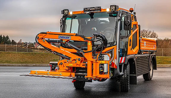 Ein orangefarbener Hansa mit einem Frontausleger an dem eine Astschere montiert ist. Das Fahrzeug steht auf einer asphaltierten Fläche und ist für Einsätze im Kommunalbereich konzipiert. Im Hintergrund sind Bäume und ein eingezäuntes Gelände sichtbar.