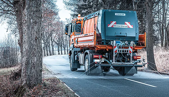 Ein orangefarbener Winterdienst-MAN fährt auf einer schmalen Straße, umgeben von Bäumen. Auf dem Fahrzeug ist ein Schild mit der Aufschrift „Winterdienst“ sichtbar. Der Lkw bringt gerade Sole auf die Straße um diese von Glatteis zu befreien.