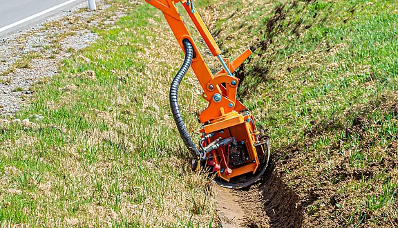Ein orangefarbene Grabenfräse an einem Frontausleger bearbeitet den Boden am Rand einer Straße. Es hat eine rotierende Schneidevorrichtung, die eine schmale Furche im Erdreich schafft. Die Umgebung zeigt frisches, grünes Gras.