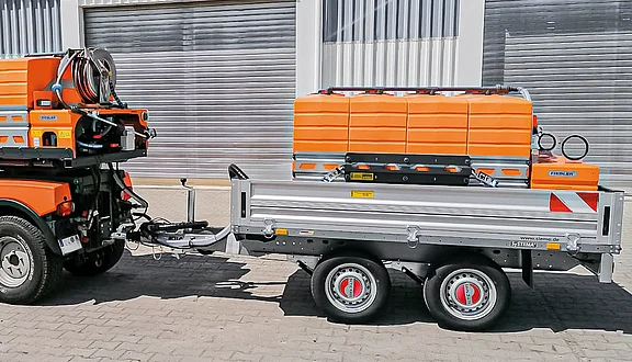 Ein orangefarbener Anhänger ist an einem Fahrzeug befestigt. Der Anhänger hat eine silberne Plattform und wird von einem orangefarbenen Transporter gezogen. Auf dem Anhänger befindet sich ein Tanksystem, das zum Gießen oder Bewässern da ist.