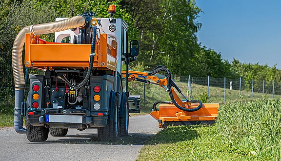 Ein Kommunalfahrzeug steht am Straßenrand. Es ist mit einem seitlichen Mähwerk ausgestattet, das zum Mähen von Gras und Unkraut dient. Am Heck befindet sich ein Gebläse, dass das Mähgut von der Fahrbahn beseitigt. Im Hintergrund sind Bäume und ein Zaun sichtbar, was auf eine ländliche Umgebung hinweist.