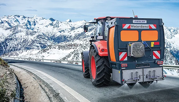 Ein roter Traktor fährt auf einer kurvenreichen Straße in den Bergen, umgeben von schneebedeckten Gipfeln. Auf der Rückseite des Traktors befindet sich die Aufschrift "Winterdienst". Der Traktor ist mit einer Solesprühanlage ausgestattet, um die Straße im Winter von Eis zu befreien.