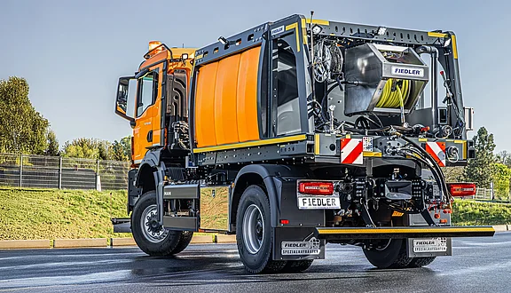 Ein orangefarbener MAN mit einem Tanksystem mit Kanalspüler, steht auf einer Straße. Der Lkw ist von der Seite sichtbar und zeigt Details wie Aufkleber und Verkleidungen. Im Hintergrund sind grüne Flächen und ein Zaun zu erkennen.