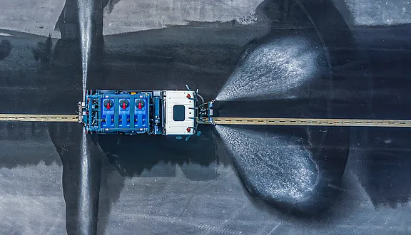 Ein MAN sprüht Wasser mit Schwemmdüsen auf eine asphaltierte Fläche. Die Wasserfontänen bilden einen breiten Schwung, während der Lkw auf einer geraden Linie fährt. Der Fokus liegt auf dem Fahrzeug und dem Wasser, das den Asphalt reinigt.