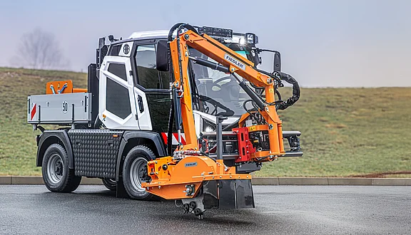 Ein Hansa Kommunalfahrzeug mit einem orangefarbenen Frontausleger. Der Ausleger ist mit einer Stubbenfräse ausgestattet, die zum entfernen von Baumstubben ist. Das Fahrzeug hat eine weiße und graue Lackierung und steht auf einer asphaltierten Fläche.