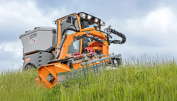 Ein kleiner orangefarbener Knicklenker mit einem großen Mähwerk an einem Frontausleger, fährt durch hohes Gras. Im Hintergrund sind graue Wolken zu sehen.