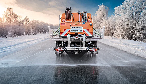 Winterdienst-Fahrzeug mit Fiedler Solesprüher-Pritschenaufbau: Ausbringung von Salzlösung zur Glatteisbekämpfung auf frostiger Straße vor verschneiter Waldkulisse für die zuverlässige Verkehrssicherung.
