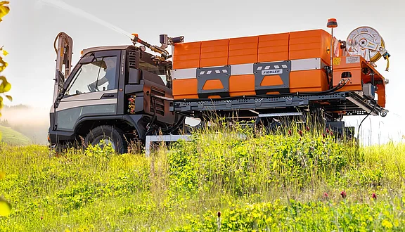 Ein Kommunalfahrzeug steht auf einer Wiese, ausgestattet mit einem orangefarbenen Tanksystem. Der Lkw hat eine robuste Bauweise und ist bereit für den Gießeinsatz in der Natur. Im Hintergrund sind sanfte Hügel und eine klare Atmosphäre zu erkennen.