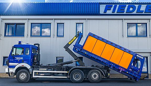 Ein blauer MAN mit einem Tanksystem auf einem Abrollcontainer, der sich anhebt, steht vor einem modernen Gebäude mit dem Schriftzug "Fiedler". Der Lkw hat eine Kranvorrichtung, die den Abrollcontainer auf sein Fahrgestell zieht.