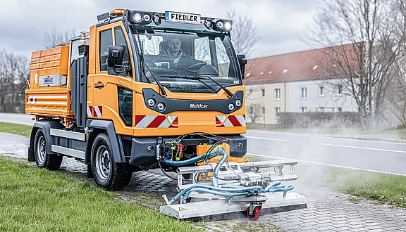 Ein orangefarbener Multicar, der mit einer Heißwasseranlage in Kombination mit einem Wildkrautbekämpfungssystem auf einem Weg fährt. Im Vordergrund sind die Anbaugeräte und ein Fußweg mit starkem Wildkrautbewuchs sichtbar, während im Hintergrund ein Wohngebäude zu sehen ist.