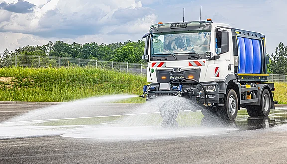 Ein MAN LKW spritzt mit Fiedler Schwemmdüsen den Schmutz von einer asphaltierten Oberfläche. Er hat einen großen Fiedler Wassertank und ist für die Straßenreinigung oder Bewässerung konzipiert. Im Hintergrund sind grüne Landschaften und ein bewölkter Himmel sichtbar.