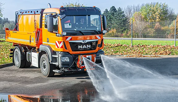 Ein orangefarbener MAN-Lkw mit einem Wassertank wird auf einer befestigten Fläche gezeigt. Der Lkw ist mit Schwemmdüsen ausgestattet, die Wasser zur Straßenreinigung versprühen. Im Hintergrund sind Bäume und ein Zaun sichtbar.