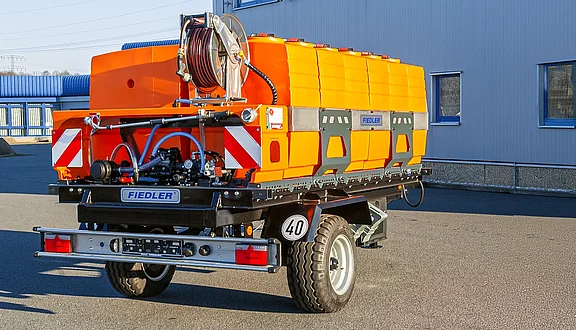 Ein orangefarbener Tankanhänger mit einem Schlauchaufroller. Im Hintergrund ist eine graue Wand und ein Gebäude sichtbar.