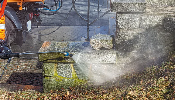 Ein Arbeiter reinigt mit einem Hochdruckreiniger einen steinernen Sockel. Wasserstrahlen entfernen grüne Algen und Schmutz von der Oberfläche. Im Hintergrund sind weitere Steine und eine gepflasterte Fläche sichtbar.