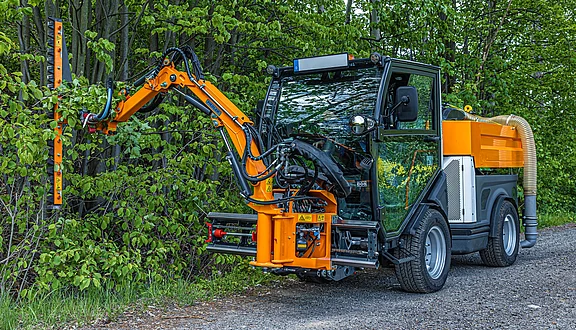 Ein orangefarbenes Kommunalfahrzeug steht am Rand einer Straße. Das Fahrzeug hat einen großen, beweglichen Frontausleger, der zum Schneiden von Hecken und kleinen Ästen genutzt wird. Im Hintergrund sind Bäume und frisches grünes Laub sichtbar.
