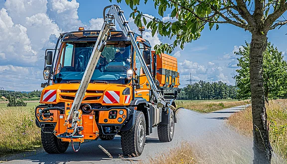 Effizientes Wassermanagement: Ein Unimog mit einem Gießarm und Tanksystem sorgt für die fachgerechte Bewässerung junger Bäume an abgelegenen Nebenwegen.