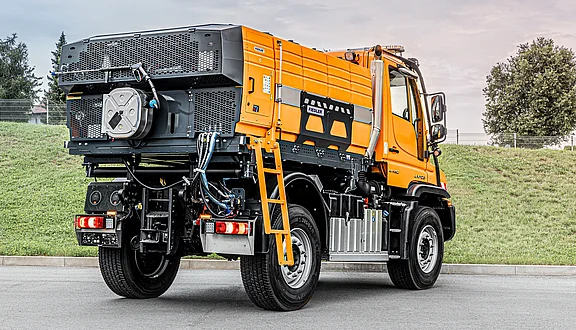 Ein orangefarbener Unimog steht auf einem gepflasterten Weg mit einer grünen Rasenfläche im Hintergrund. Die Rückseite des Fahrzeugs zeigt eine große, aufgesetzte Heißwasseranlage und verschiedene Anschlüsse sowie eine Leiter zum klappen an der Seite.