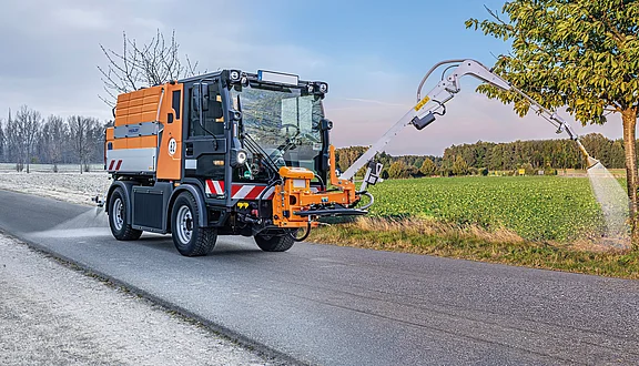 Ein Citymaster mit einem Gießarm, der einen Baum Gießt, fährt entlang einer Straße. Am Heck hat er eine Solesprühanlage um im Winter Glätte zu bekämpfen. Im Hintergrund sind Felder und Bäume zu sehen. Das soll die Flexibilität des Tanksystems darstellen - Sommer und Winternutzung.