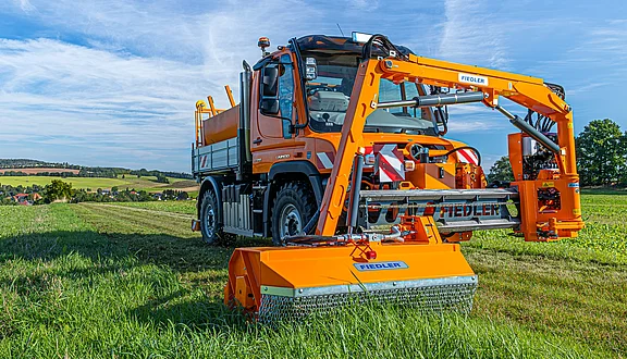 Ein orangefarbener Unimog steht auf einem Feld. Er ist mit einem großen Frontausleger ausgestattet, der zum Mähen dient. Im Hintergrund sind sanfte Hügel und Felder zu sehen.