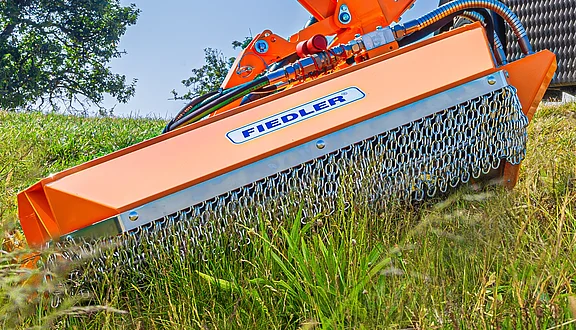 Das Bild zeigt einen orangefarbenen Mähkopf an einem Frontausleger, der im Gras steht. Die Marke "FIEDLER" ist deutlich sichtbar. Im Hintergrund sind Bäume und Wiesen zu erkennen.