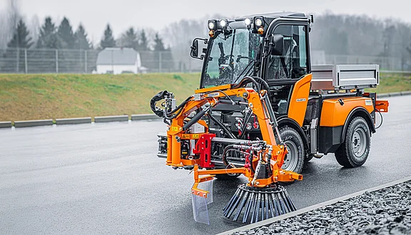 Ein Kärcher Kommunalfahrzeug, ausgestattet mit einer Wildkrautbürste an einem Frontausleger, steht auf einer nassen Straße. Im Hintergrund sind Bäume und eine gepflegte Landschaft sichtbar. Das Fahrzeug ist bereit für die Beseitigung von Wildkraut an Straßenrändern und Fußwegen.