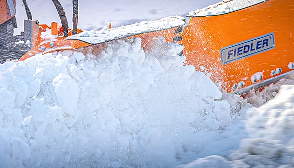 Eine orangefarbene Schneefräse entfernt Schnee. Der Schnee wird in einer großen weißen Wolke aufgewirbelt, während die Fräse in Bewegung ist. Die Maschinenmarke "FIEDLER" ist deutlich sichtbar.