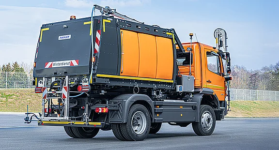 Ein oranges, robustes Kommunalfahrzeug steht einsatzbereit auf einer asphaltierten Straße. Es ist mit einem silbernen Fiedler-Gießarm an der Front und einem großen Fiedler-Tanksystem auf der Ladefläche ausgestattet, konzipiert für die effiziente mobile Bewässerung im städtischen und ländlichen Raum.