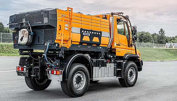 Ein orangefarbener Unimog steht auf einer Straße. Er hat ein großes Tanksystem geladen, das auch zur Heißwasserausbringung genutzt werden kann. Der Unimog ist robust und für schwere Lasten ausgelegt. Im Hintergrund sind Bäume und eine Grünfläche sichtbar.