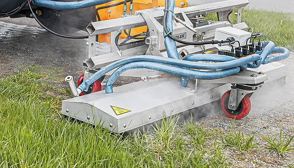 Ein Gerät zur Unkrautbekämpfung spritzt Dampf auf eine Grasfläche. Die Maschine hat verschiedene Schläuche und Räder. Die Umgebung zeigt eine Mischung aus befestigtem Boden und Rasen, wobei der Dampfausstoß deutlich sichtbar ist.