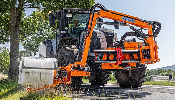 Ein Traktor mit einem orangefarbenen Frontausleger zur Straßenreinigung fährt entlang einer Straße. Das Gerät ist mit einer Bürste ausgestattet, die die Leitpfosten sauber hält. Bäume säumen die Straße, und die Sonne scheint.