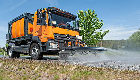 Ein orangefarbener Mercedes-Lkw sprüht Wasser auf eine Straße. Er hat einen großen Wassertank hinten und einen breiten Schwemmbalken vorne. Im Hintergrund sind Bäume und eine grüne Wiese sichtbar. Die Szene vermittelt eine Reinigung der Straße.