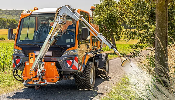Ein AEBI VT Kommunalfahrzeug am Rand einer Landstraße; der Gießarm ist über einer Pflanzung positioniert. Im Hintergrund erstreckt sich eine ländliche Szenerie mit grünen Feldern und Bäumen.
