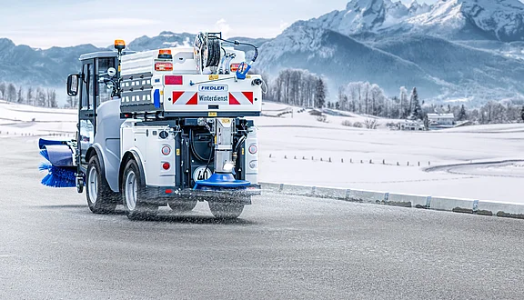 Emissionsfreier Winterdienst: Citymaster ZE mit Fiedler Winterdiensttechnik im Einsatz vor Bergkulisse. Die vollelektrische Kehrmaschine nutzt einen Frontbesen und eine aktive Solesprühanlage zur Glatteisbekämpfung auf verschneiten Straßen.