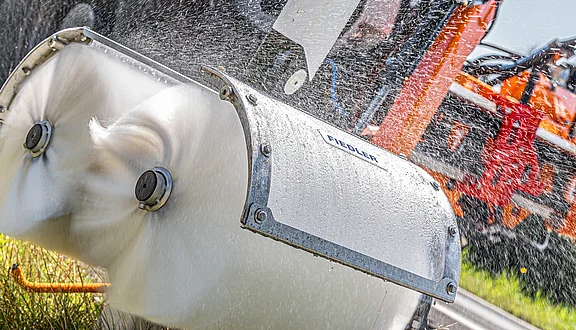 Eine Leitpfostenwaschbürste beim Reinigen eines Leitpfostens. Die rotierenden Düsen erzeugen Wasserspritzer, die den Eindruck von Kraft und Dynamik vermittelt. Im Hintergrund ist ein Traktor mit Frontausleger zu erkennen.