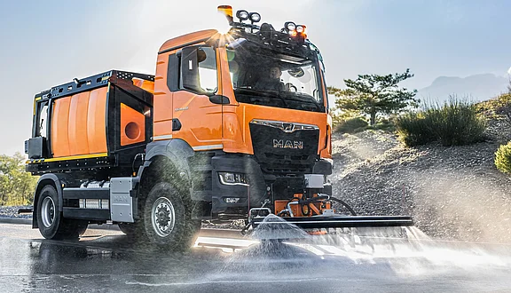 Ein oranger MAN steht auf einer Straße. Er hat ein großes Tanksystem hinten und ein Schwemmbalken an der Vorderseite, der Wasser sprüht. Im Hintergrund sind einige Bäume und Hügel sichtbar. Das Fahrzeug scheint gerade im Einsatz zu sein.