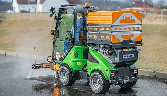 Ein Egholm mit einem orangefarbenen Tanksystem reinigt mit einem Schwemmbalken eine asphaltierte Fläche. Der Fahrer sitzt im Fahrzeug, während es in eine rechte Richtung fährt. Im Hintergrund sind unscharfe Gebäude und ein blauer Himmel sichtbar.