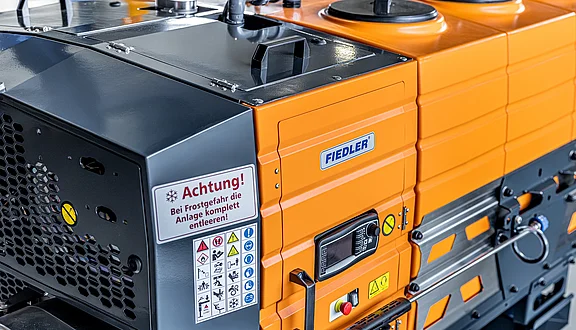 Die elektrisch angetriebene Heißwasseranlage FTS Kombipower Light E der Firma FIEDLER. Sie ist in ein oranges Tanksystem integriert. Auf der Vorderseite befinden sich Warnhinweise in deutscher Sprache. Die Maschine zeigt verschiedene Bedienelemente und Sicherheitshinweise.