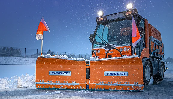 Ein orangefarbener Schneeräumer steht auf einer verschneiten Straße. Das Fahrzeug hat ein Keilpflug in gerader Stellung vorne dran, das mit reflektierenden Flaggen gekennzeichnet ist. Im Hintergrund fällt leichter Schneefall, und die Umgebung ist winterlich und ruhig.
